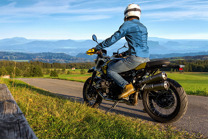 Person mit weißem Helm, blauer Jacke und gelben Handschuhen auf schwarzem Motorrad mit Berglandschaft im Hintergrund.