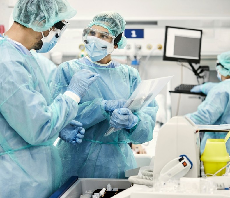 Foto von einer Ärztin und einem Arzt, die in Schutzkleidung auf einer Intensivstation stehen und sich beratschlagen. Foto von einer Ärztin und einem Arzt, die in Schutzkleidung auf einer Intensivstation stehen und sich beratschlagen.