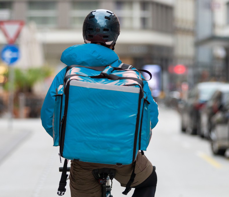 Die Person trägt einen dunklen Helm, sowie eine Jacke und einen Lieferdienst-Rucksack im gleichen hellblauen Farbton. Die Person trägt einen dunklen Helm, sowie eine Jacke und einen Lieferdienst-Rucksack im gleichen hellblauen Farbton.