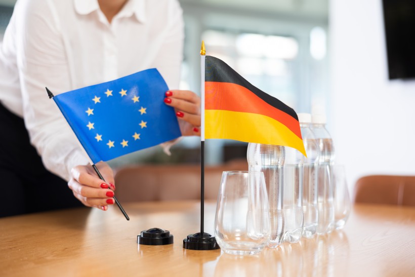 Eine Person im weißen Hemd mit rot lackierten Fingernägeln stellt zwei kleine Tischflaggen auf einem Konferenztisch auf: eine blaue Flagge der Europäischen Union mit gelben Sternen und eine schwarz-rot-goldene Flagge Deutschlands. Daneben stehen mehrere Wasserflaschen und leere Gläser.