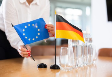 Eine Person im weißen Hemd mit rot lackierten Fingernägeln stellt zwei kleine Tischflaggen auf einem Konferenztisch auf: eine blaue Flagge der Europäischen Union mit gelben Sternen und eine schwarz-rot-goldene Flagge Deutschlands. Daneben stehen mehrere Wasserflaschen und leere Gläser. Eine Person im weißen Hemd mit rot lackierten Fingernägeln stellt zwei kleine Tischflaggen auf einem Konferenztisch auf: eine blaue Flagge der Europäischen Union mit gelben Sternen und eine schwarz-rot-goldene Flagge Deutschlands. Daneben stehen mehrere Wasserflaschen und leere Gläser.